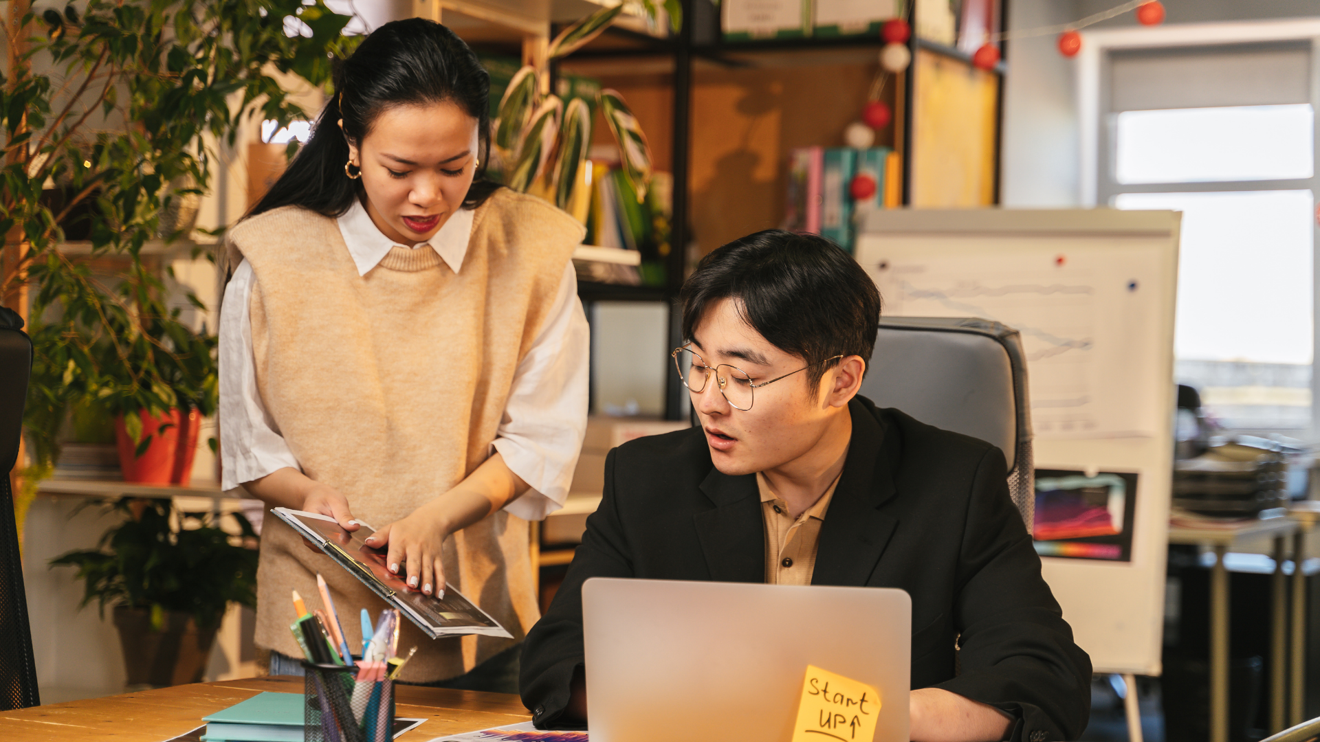 An Asian lady talking to an Asian man who is seated on a desk with an open laptop